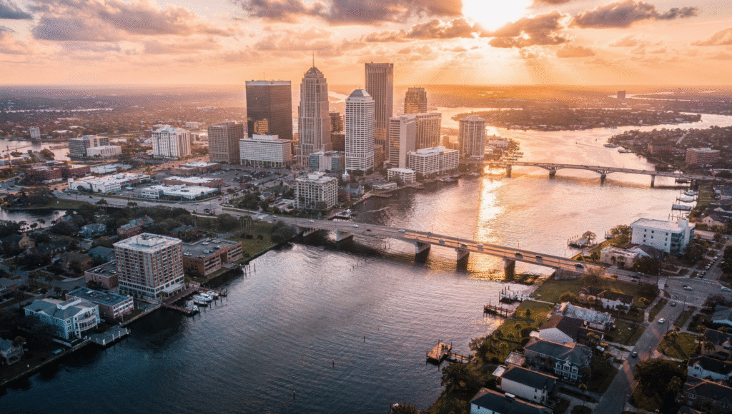 Wide aerial view of downtown Jacksonville Florida with the St. Johns River, bridges, and nearby neighborhoods at sunset, bright sky and reflective water, high quality real estate photography